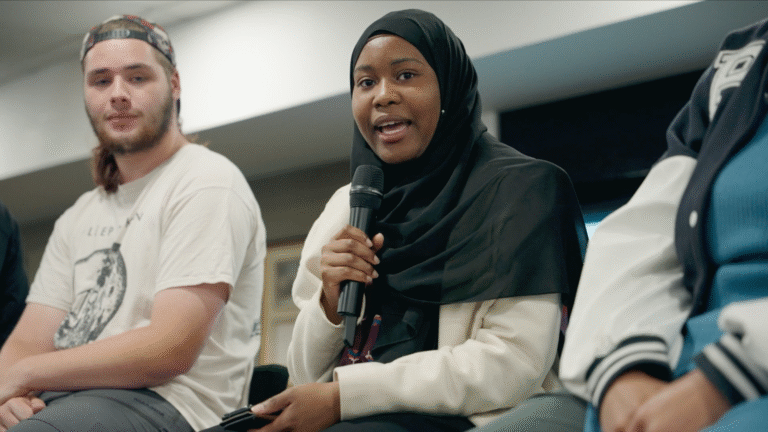 Ayyat Adiga=un holding a microphone talking at the Sandwell Story launch. She is sat on a chair and next to her is Brandon. They are both in white, she has a black head scarf on.