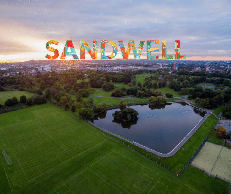 Aerial shot of Sandwell with the sun setting in the distance, you can see water, grass and trees. On the horizon is a Sandwell Story branded outline of 'Sandwell'