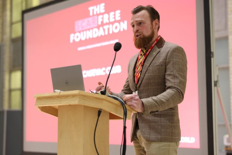 Karl speaking at a lecturn in front of a pink Scar Free Foundation screen. He has a beard, is wearing a brown suit jacket and is speaking into a microphone